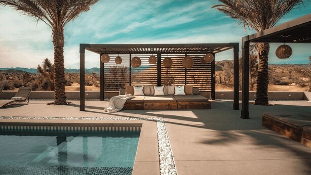 A pool area with a wooden deck, a seating area with cushions, and a pergola with hanging lights. - Iconic 6BR Desert Retreat w/ Pool, Pickleball & Fire Pit Joshua Tree Mansion by Avantstay (Joshua Tree)