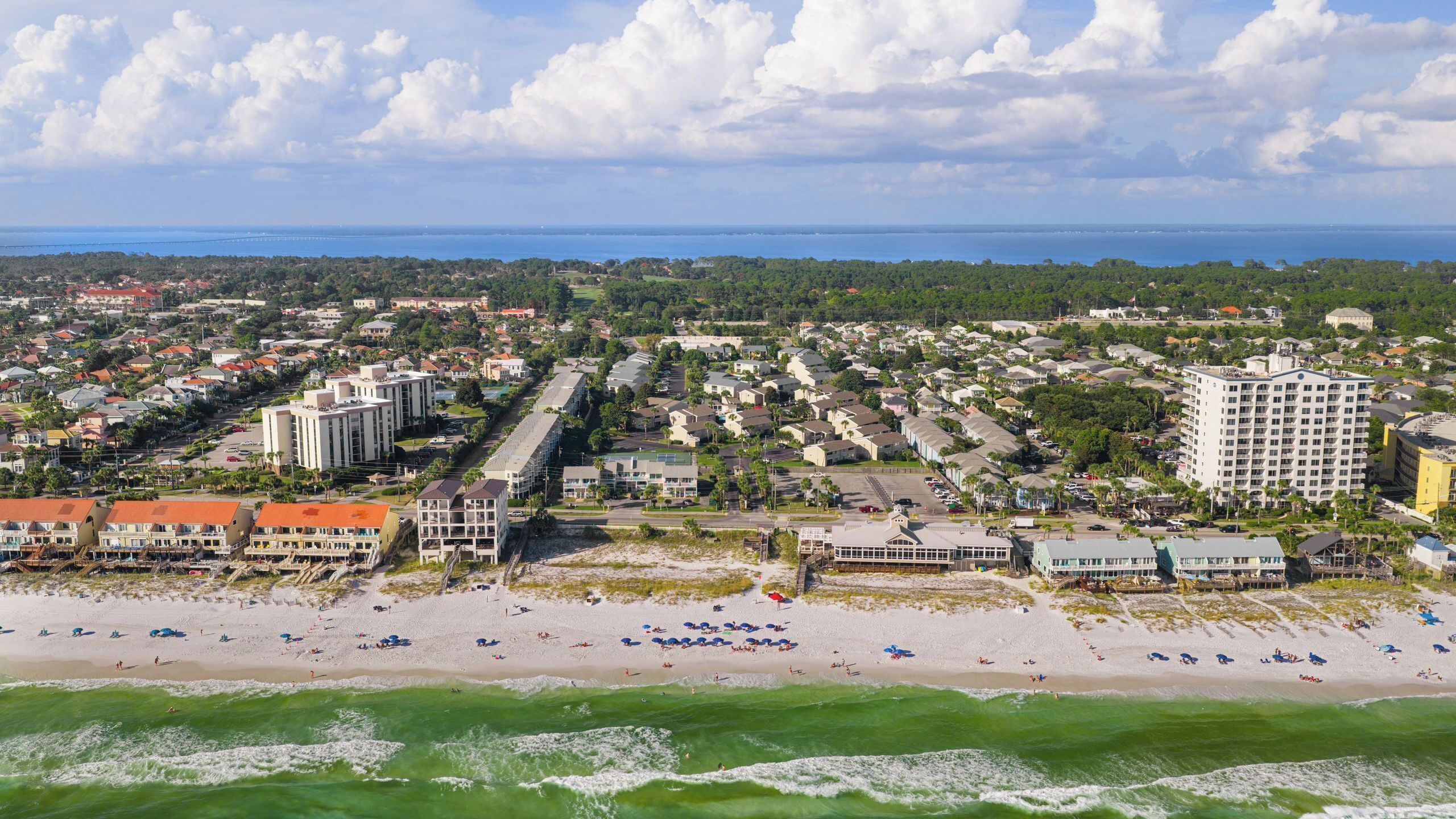 An aerial view of a coastal town with beachfront hotels, a marina, and a bridge in the distance.