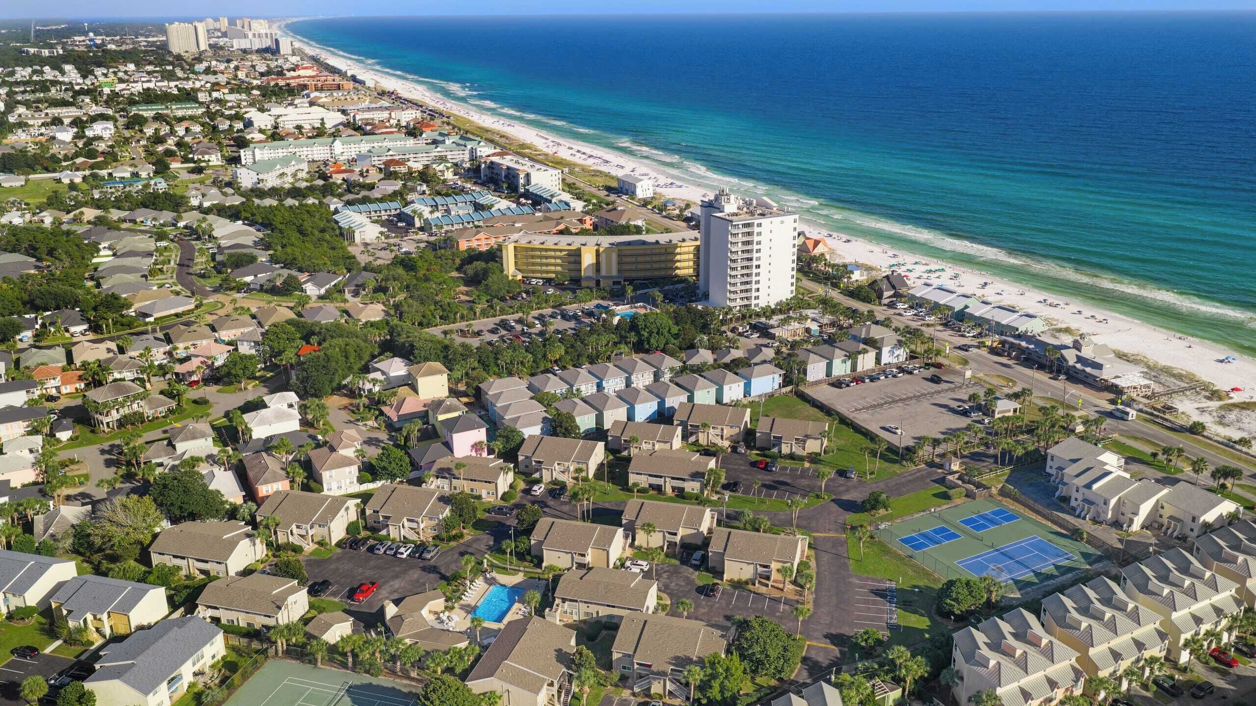 Vue aérienne d’un quartier résidentiel situé sur la côte, avec une plage, un grand complexe hôtelier et un court de tennis.