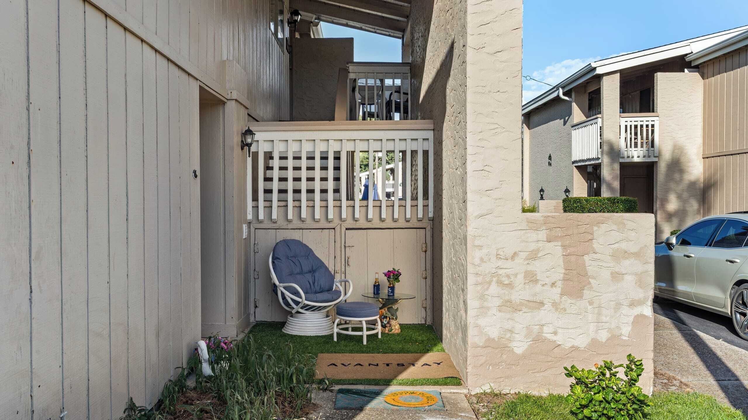 A residential building with a gated entrance, a small outdoor seating area with a chair and table, and a doormat with the text "AVANTSTAY".