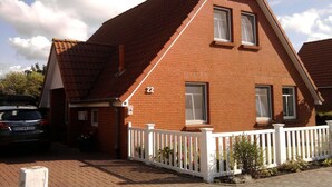 A two-story brick house with a white picket fence, a car parked in the driveway, and a clear sky. - Holiday home with 3 bedrooms (Greetsiel)