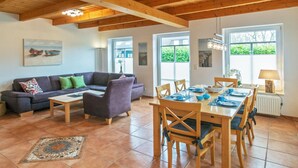 A spacious living area with a wooden dining table, chairs, and a sofa. There are large windows providing natural light and views of greenery outside. - Holiday home with 3 bedrooms (Greetsiel)