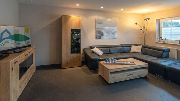 Een moderne woonkamer met een hoekbank, een houten kast met een televisie en een groot raam met zonwering.