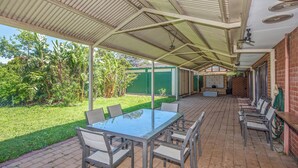 Une terrasse extérieure avec une table et des chaises à plateau en verre, entourée d’une zone abritée dotée d’un toit en métal et d’un sol en briques.