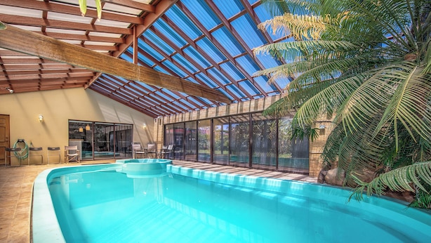 Une piscine intérieure dotée d’un toit en verre, entourée de palmiers et agrémentée d’un espace patio.