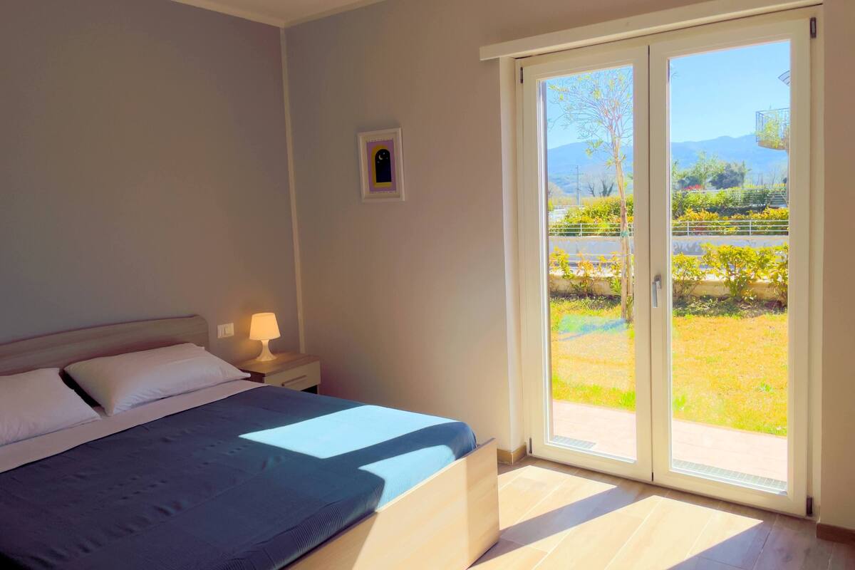 Une chambre à coucher avec un lit, une table de chevet avec une lampe, un petit tableau encadré et une porte-fenêtre coulissante donnant sur un balcon avec vue sur la verdure et les montagnes.