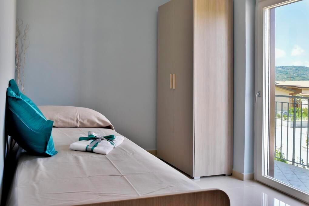 Een netjes opgemaakt bed met een blauwgroen kussen en een witte handdoek, een houten garderobe en een balkon met uitzicht.