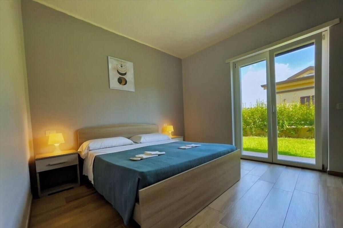 Une chambre à coucher comprenant un lit, des lampes de chevet, une table de nuit, une porte-fenêtre coulissante donnant sur un jardin et un tableau encadré au mur.