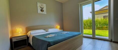A bedroom with a bed, bedside lamps, a nightstand, a sliding glass door to a garden, and a framed picture on the wall.