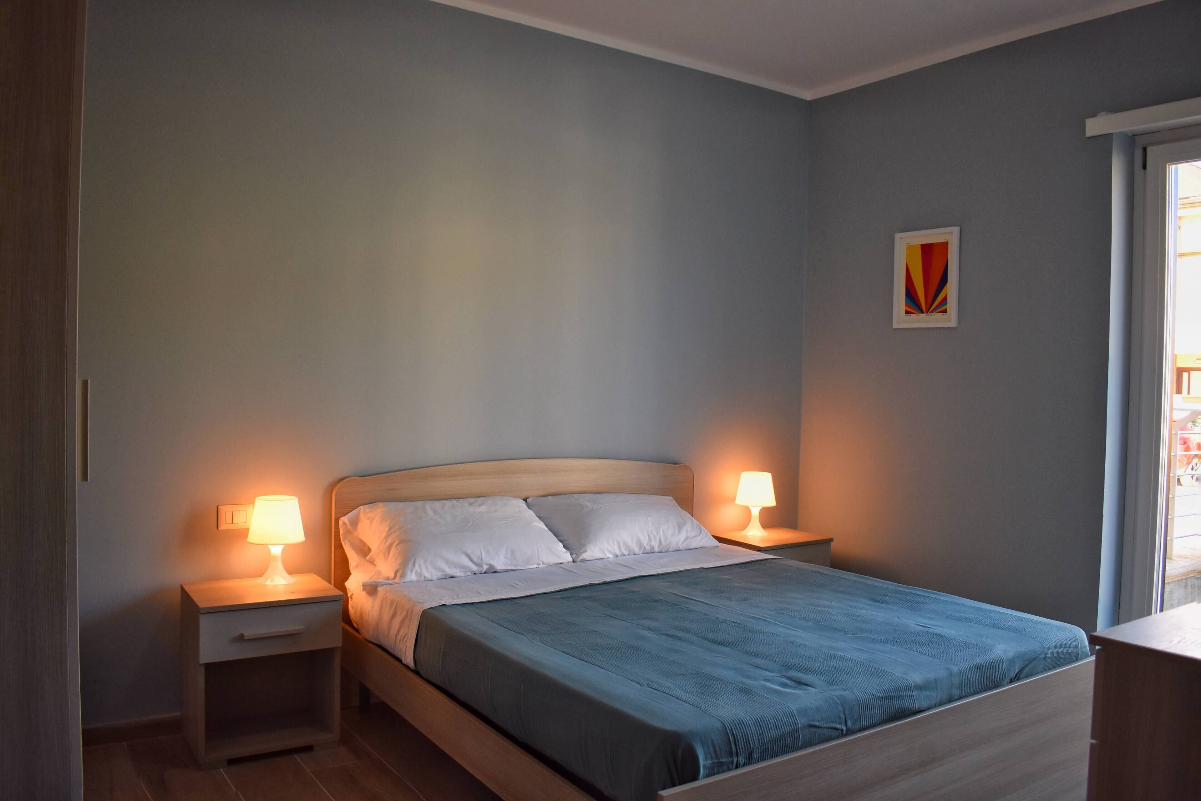 Una cama bien hecha con colcha azul y almohadas blancas, flanqueada por dos mesitas de noche con lámparas, en una habitación con paredes de tonos claros y una pequeña obra de arte.