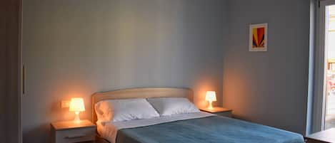Una cama bien hecha con colcha azul y almohadas blancas, flanqueada por dos mesitas de noche con lámparas, en una habitación con paredes de tonos claros y una pequeña obra de arte.