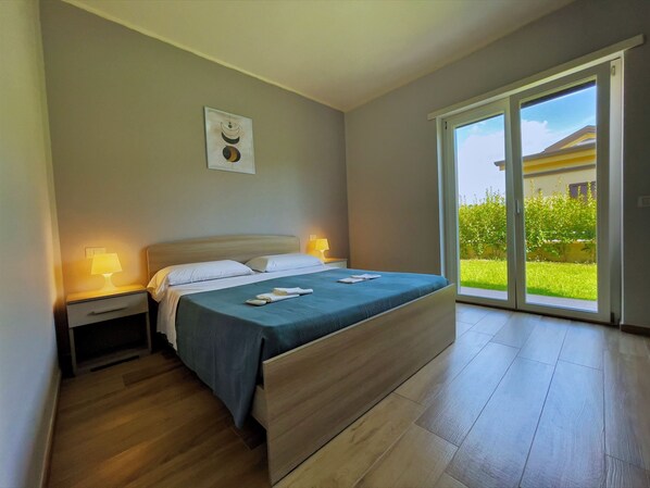 Una camera da letto moderna con un letto grande, due abat-jour, un comodino e una porta scorrevole in vetro che dà su un giardino.