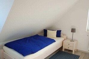 Une petite chambre avec un lit simple, une couverture bleue, une lampe de chevet et un petit tapis.
