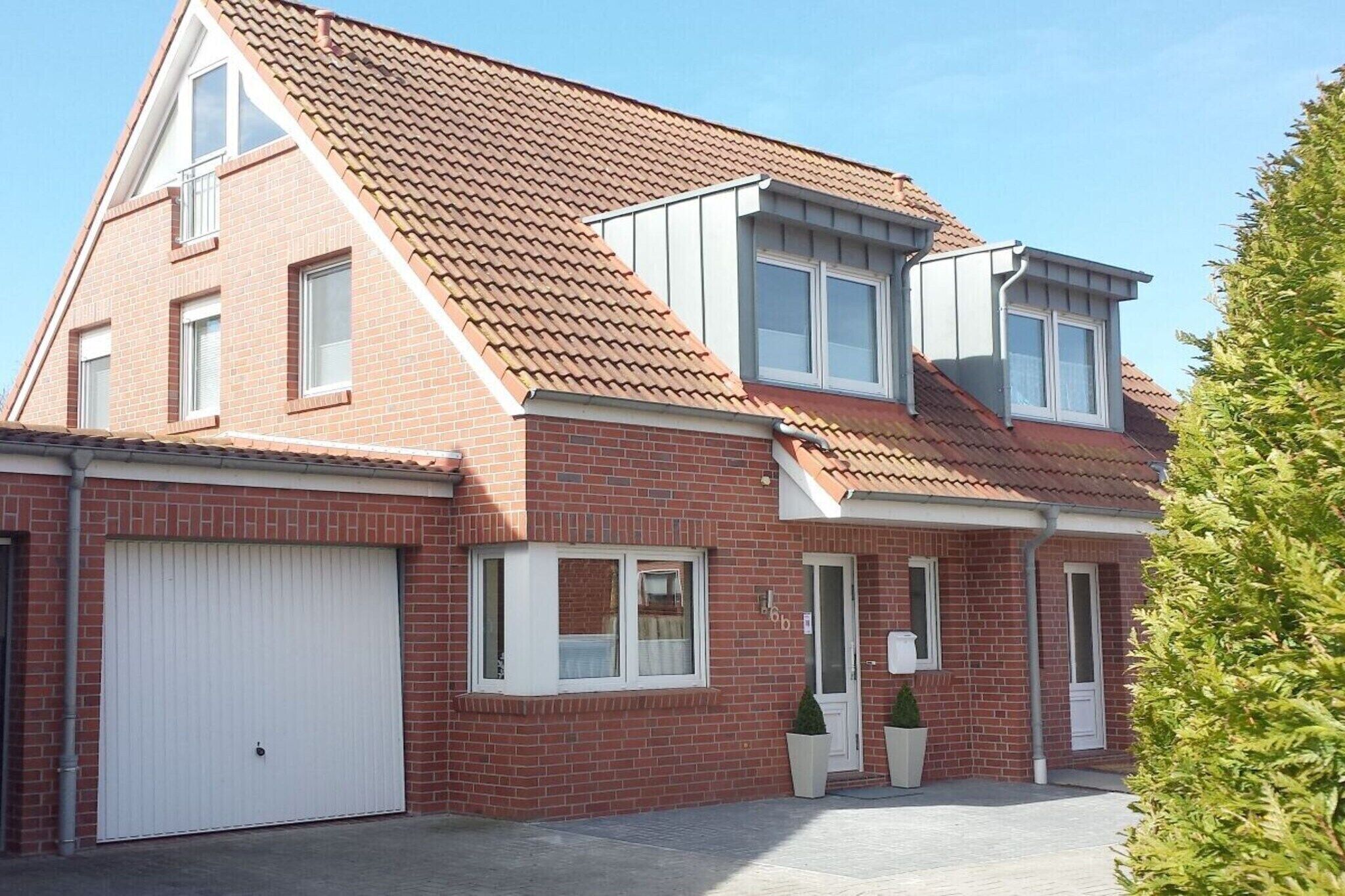 Une maison à deux étages avec une façade en briques rouges, une porte de garage blanche et un petit porche à l’avant, agrémenté d’un pot de fleurs.