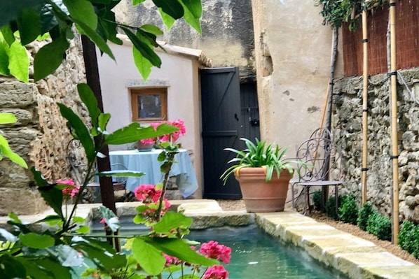 A narrow courtyard with a water feature, a table with a blue tablecloth, and potted plants.