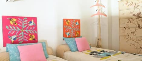 A bedroom with two beds, colorful artwork, a pink hanger, and a dresser with floral patterns.