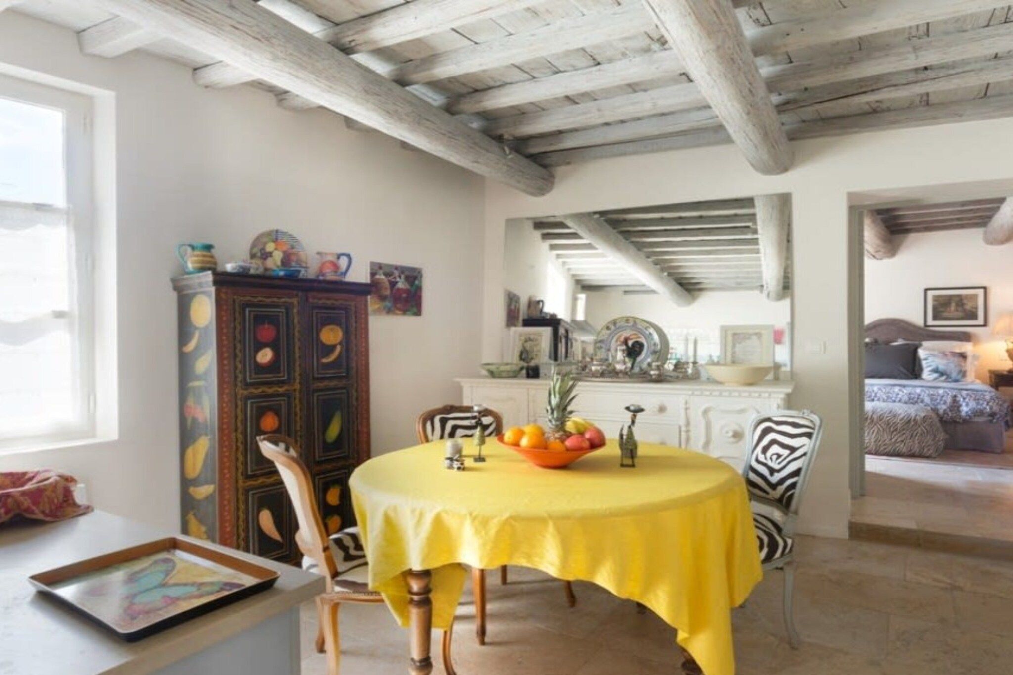 A dining area with a round table, chairs, and a yellow tablecloth. There is a cabinet with fruit paintings and a window with curtains.
