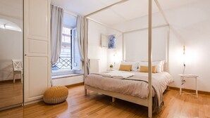 Ein Schlafzimmer mit Himmelbett, einem Fenster mit Vorhängen, einem Heizkörper und einem kleinen Nachttisch.