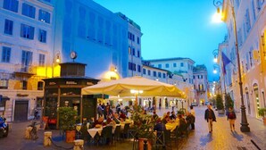 Ein Straßencafé mit Tischen und Stühlen, umgeben von Gebäuden mit Balkonen und Fenstern.