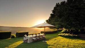 An outdoor dining setup with a table and chairs under a large umbrella, surrounded by well-manicured hedges and trees, with a scenic view in the background.
