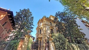 Un bâtiment historique à l’architecture riche et ornée, entouré d’arbres et agrémenté d’une grille en fer forgé.