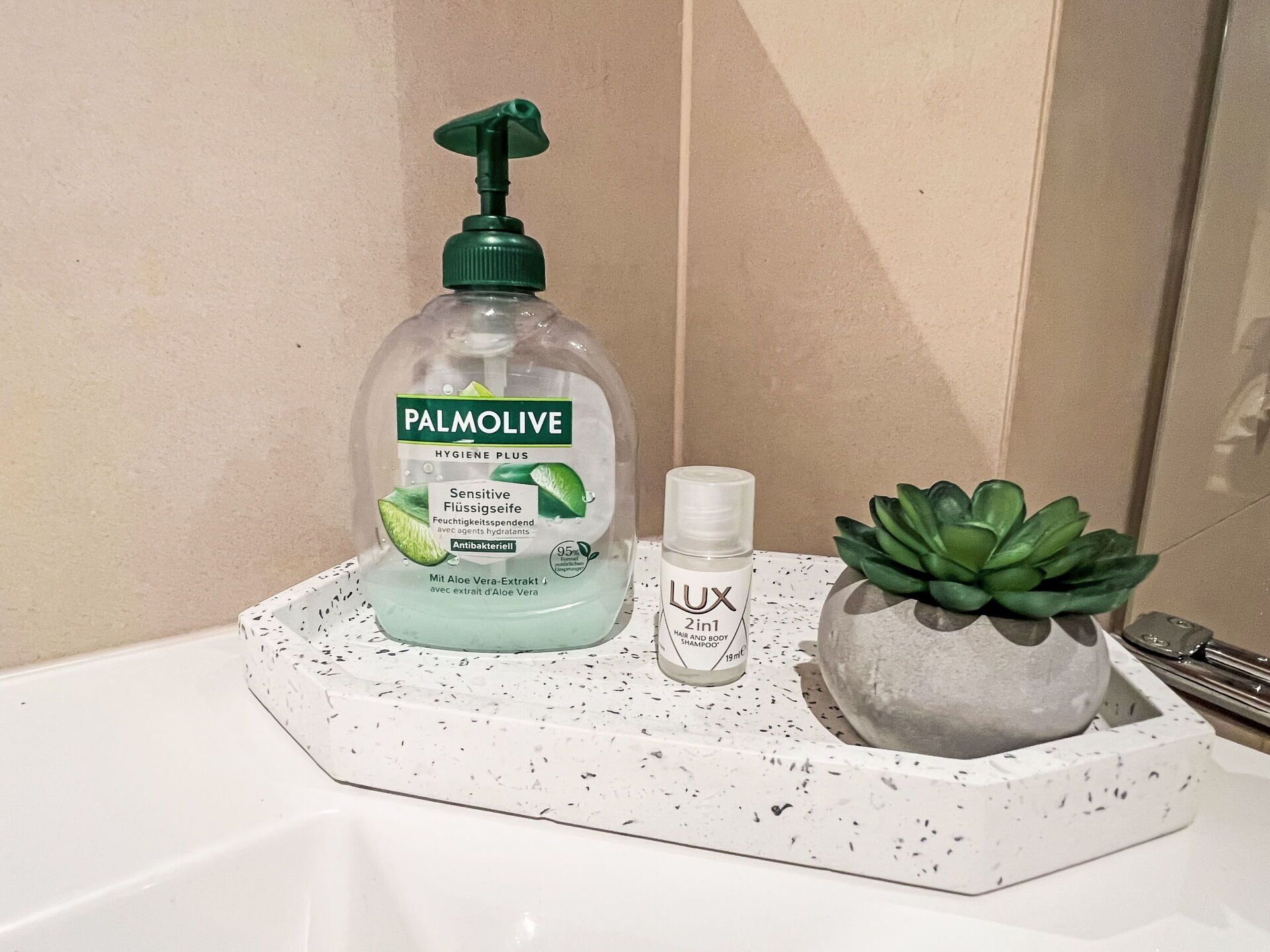 Ein Badezimmerwaschbecken mit einer Arbeitsplatte mit weißen Tupfen. Auf der Arbeitsplatte stehen eine Flasche Palmolive Handseife, eine Flasche Lux Ha- und Körper-Shampoo sowie eine kleine, in einem Topf stehende Sukkulente.