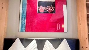 Une chambre avec un lit doté d’une tête de lit noire et de deux oreillers blancs. Au-dessus du lit, on aperçoit une œuvre d’art encadrée avec un fond rouge, et une fenêtre laissant entrevoir des fleurs roses.