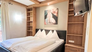 A double bed with white linens, a headboard, and a painting above it. There is a TV mounted on the wall and a wooden shelf unit to the right.