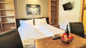 Cama bem arrumada com lençóis brancos, cabeceira de madeira e um quadro na parede. Há uma pequena mesa com uma tigela de frutas e uma garrafa.