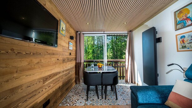 A cozy room with wooden walls, a large window, a TV, a table with chairs, and a blue sofa. - Wooden apartment with balcony (Allos)