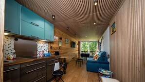 A compact kitchen with wooden cabinets, a dining area with a blue sofa, and a view of greenery outside. - Wooden apartment with balcony (Allos)