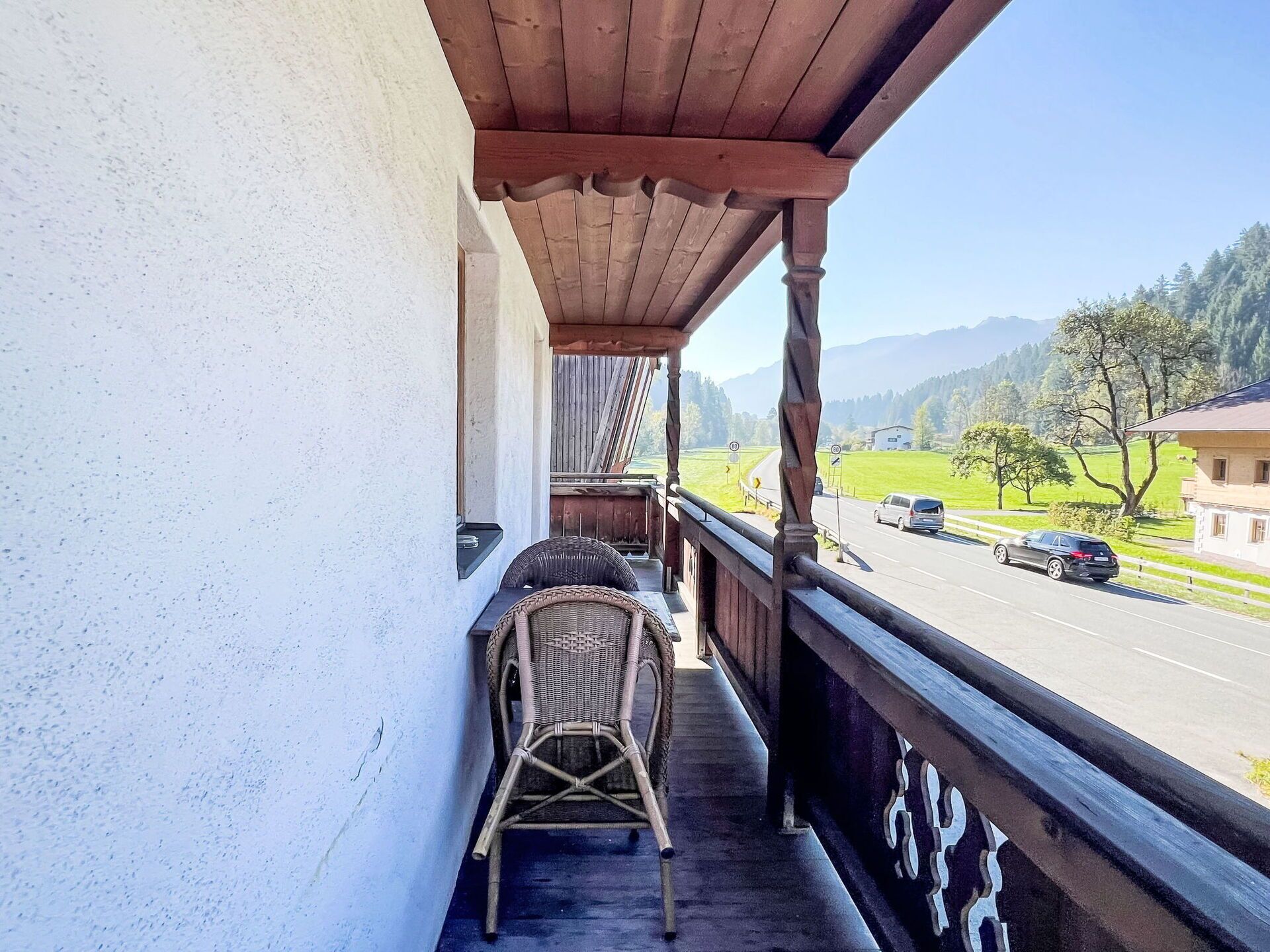 A balcony with wicker chairs, a wooden railing, and a view of a road and green fields.