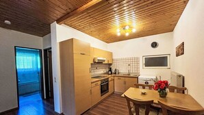 A compact kitchen with wooden cabinets, a dining table with chairs, a clock on the wall, and a window with curtains. - Nice apartment for 4 guests with WIFI, TV and balcony (Ludmannsdorf)