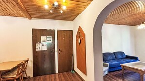 A room with wooden flooring, a dining table with chairs, a blue sofa, and a small table. There are two wooden doors with a bulletin board and a ceiling fan. - Nice apartment for 4 guests with WIFI, TV and balcony (Ludmannsdorf)