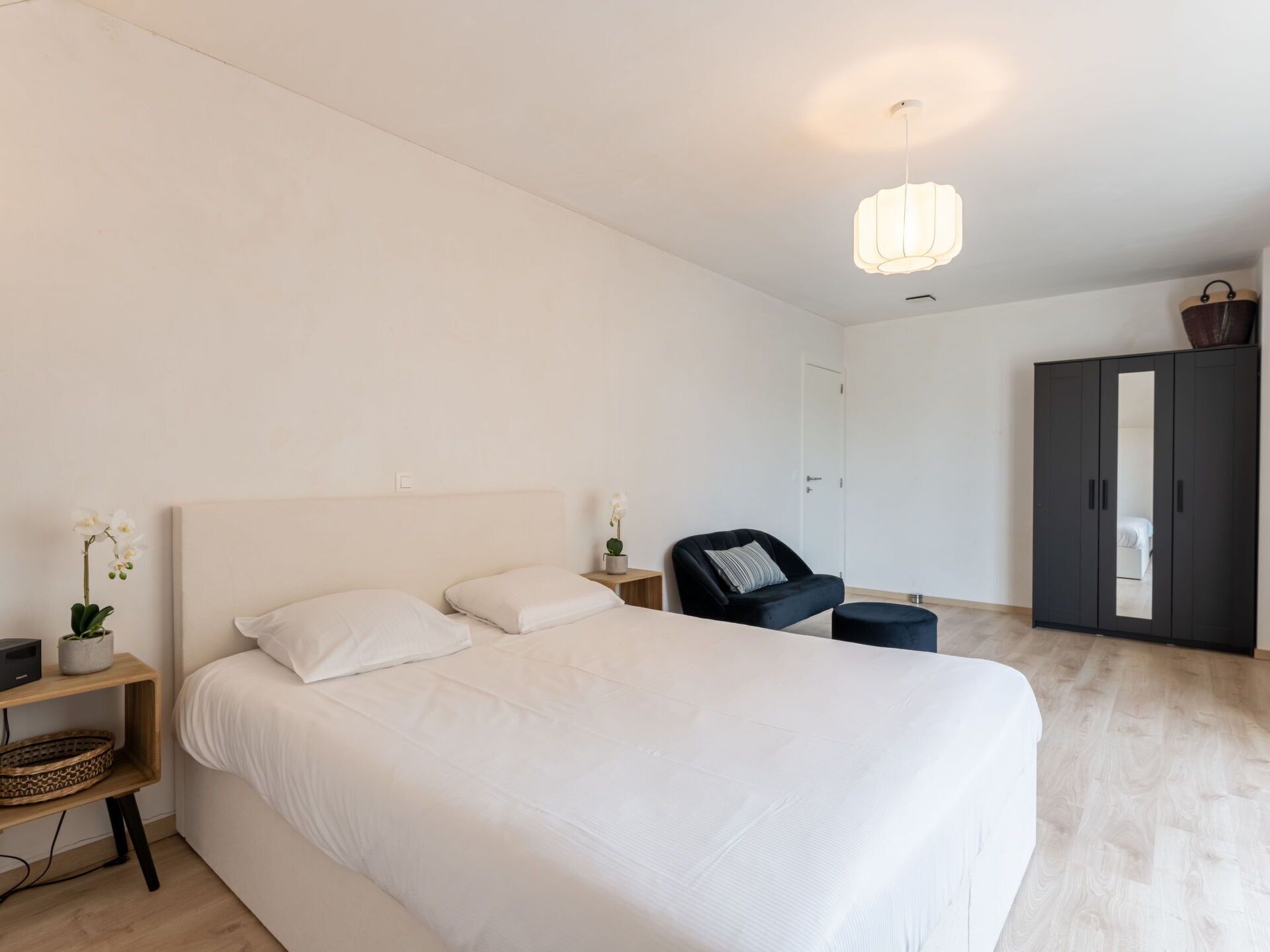 A modern bedroom with a large bed, a bedside table with a lamp, a chair, a wardrobe, and a small plant.