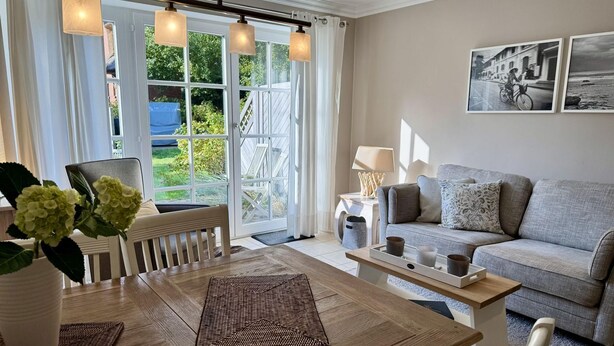 A cozy living room with a wooden dining table, a grey sofa, and a large window with curtains. - Gaathüs - Stylish vacation home under thatch in Keitum (Sylt-Ost)