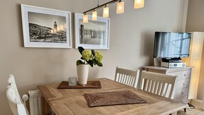 A dining area with a wooden table, white chairs, a vase with flowers, and a television on a stand. - Gaathüs - Stylish vacation home under thatch in Keitum (Sylt-Ost)
