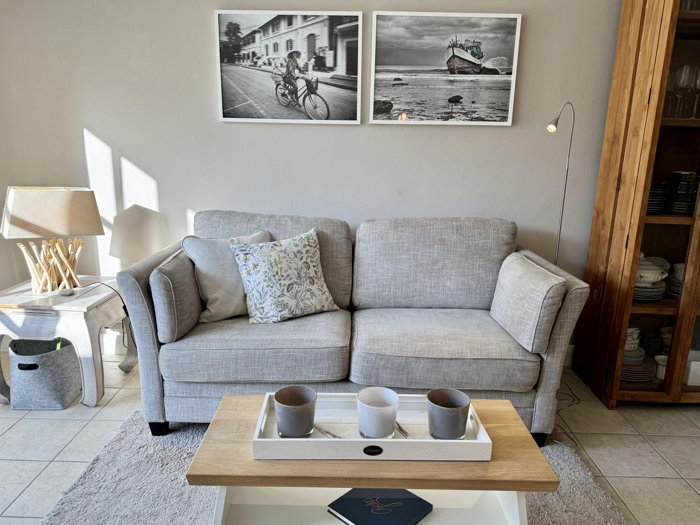 Un salon avec un canapé gris, une table basse sur laquelle se trouve un plateau avec des tasses, et deux photographies en noir et blanc accrochées au mur.