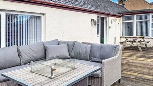 Un espace extérieur moderne, aménagé avec un canapé gris et une table en bois, situé devant une maison dont le toit est recouvert de tuiles et qui possède une cheminée visible.