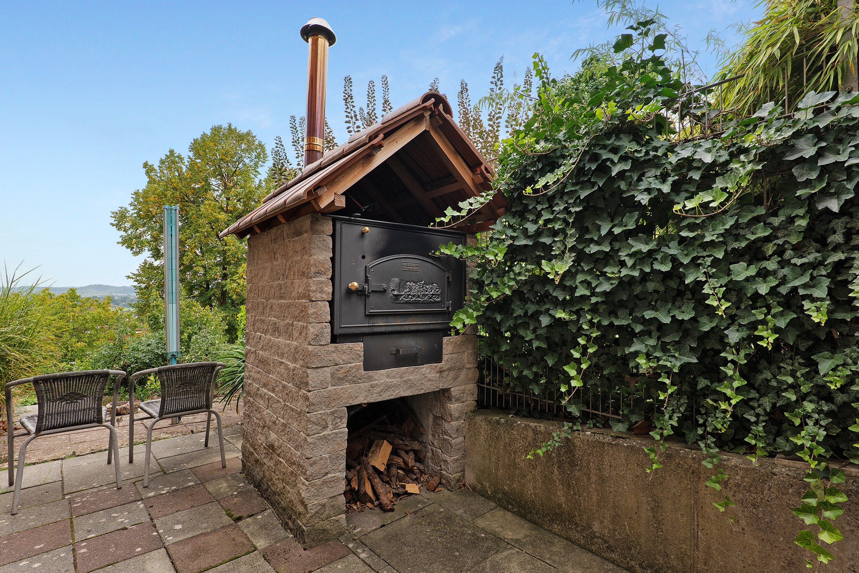 Una chimenea de piedra al aire libre con una puerta metálica, rodeada de follaje y un banco.