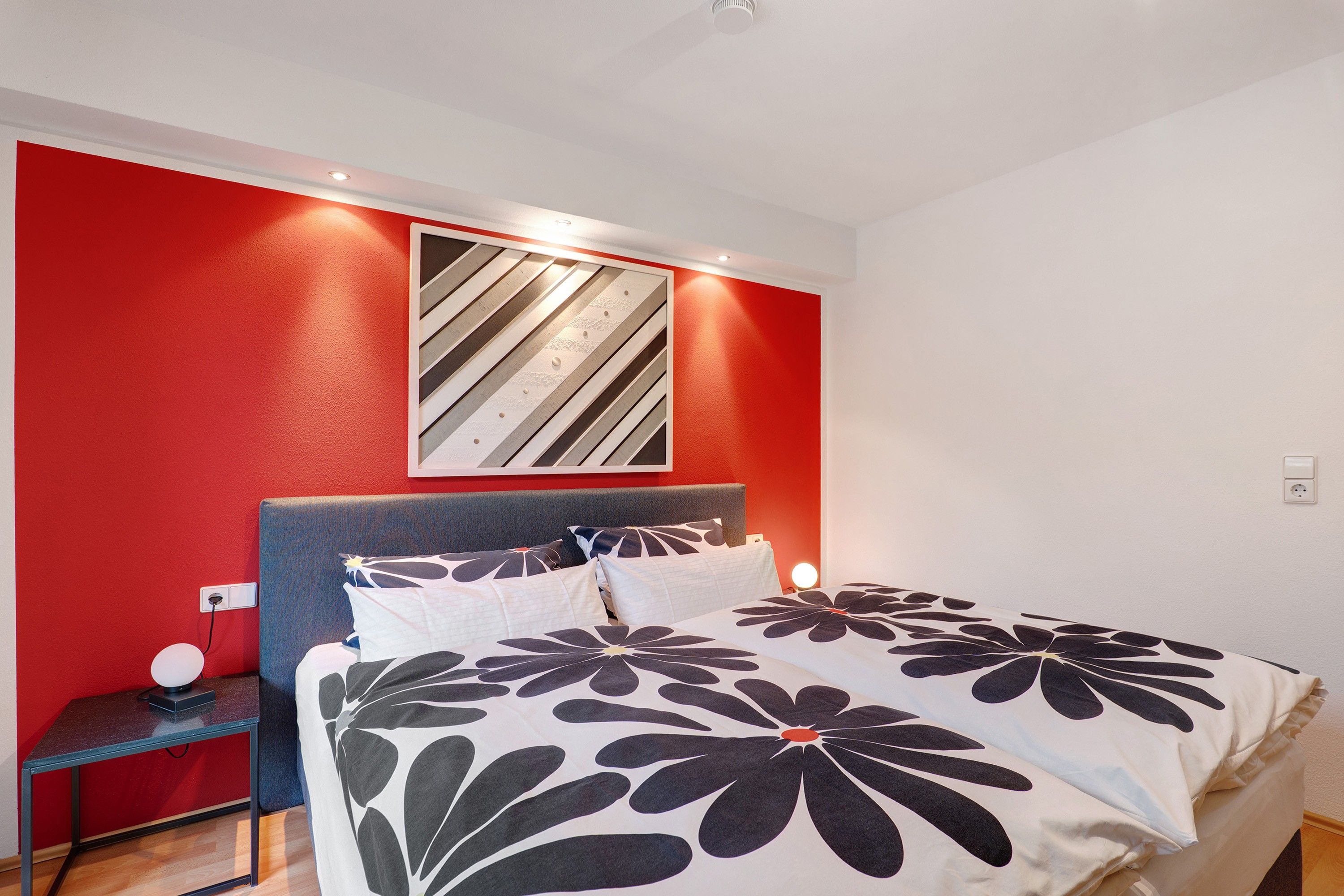 Een slaapkamer met een bed, nachtkastje, wandlamp en een rode accentmuur.
