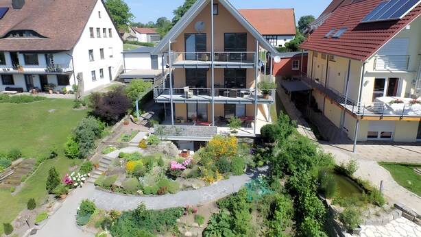 A multi-story building with a balcony, a garden with various plants and flowers, and a small pond. - Holiday Apartment Bär, 59 sqm, 1 Bedroom, Living and Dining Area, max. 3 Persons (Rosengarten)