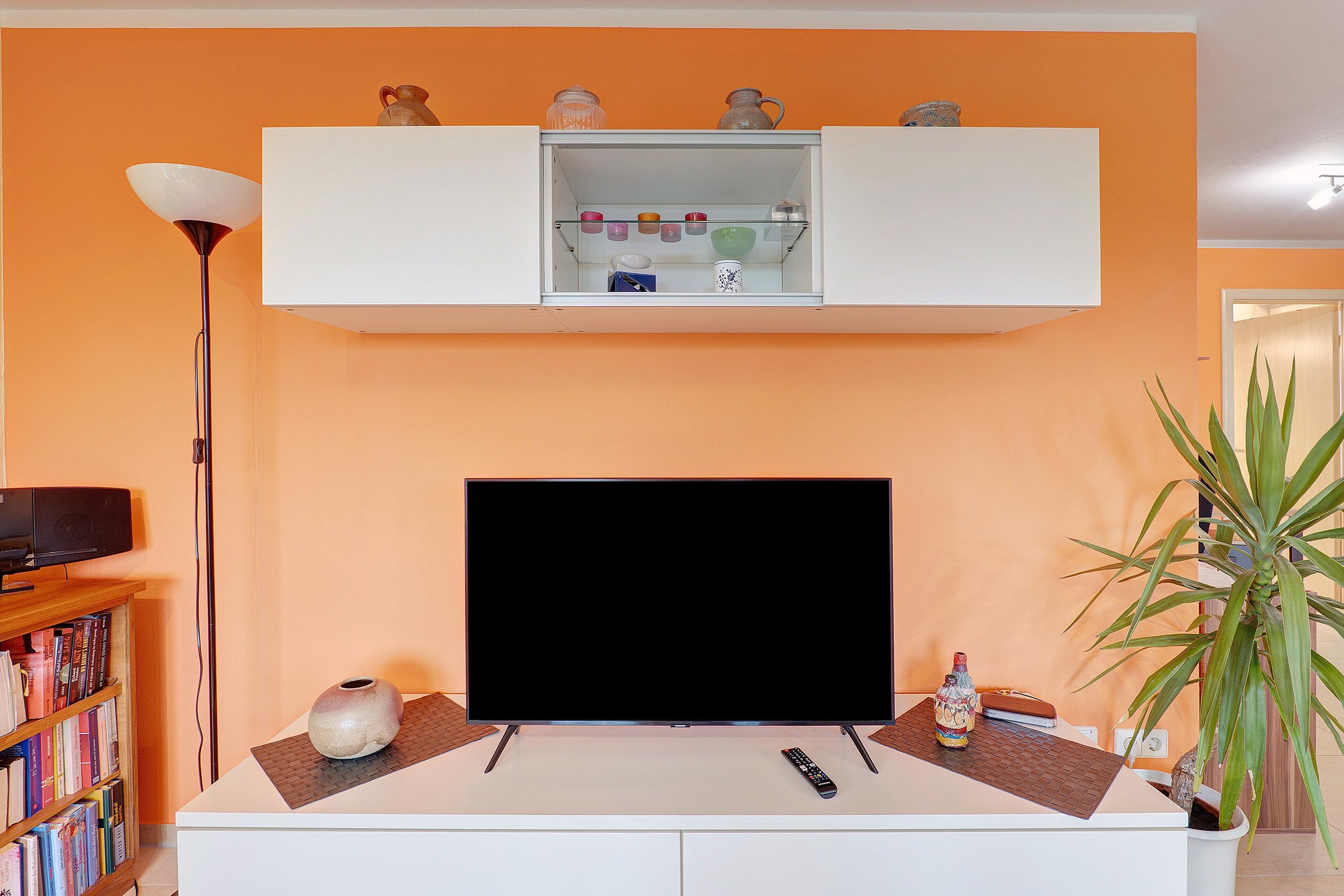 Una sala de estar con una televisión de pantalla plana, una estantería, una lámpara de pie y una planta en maceta.