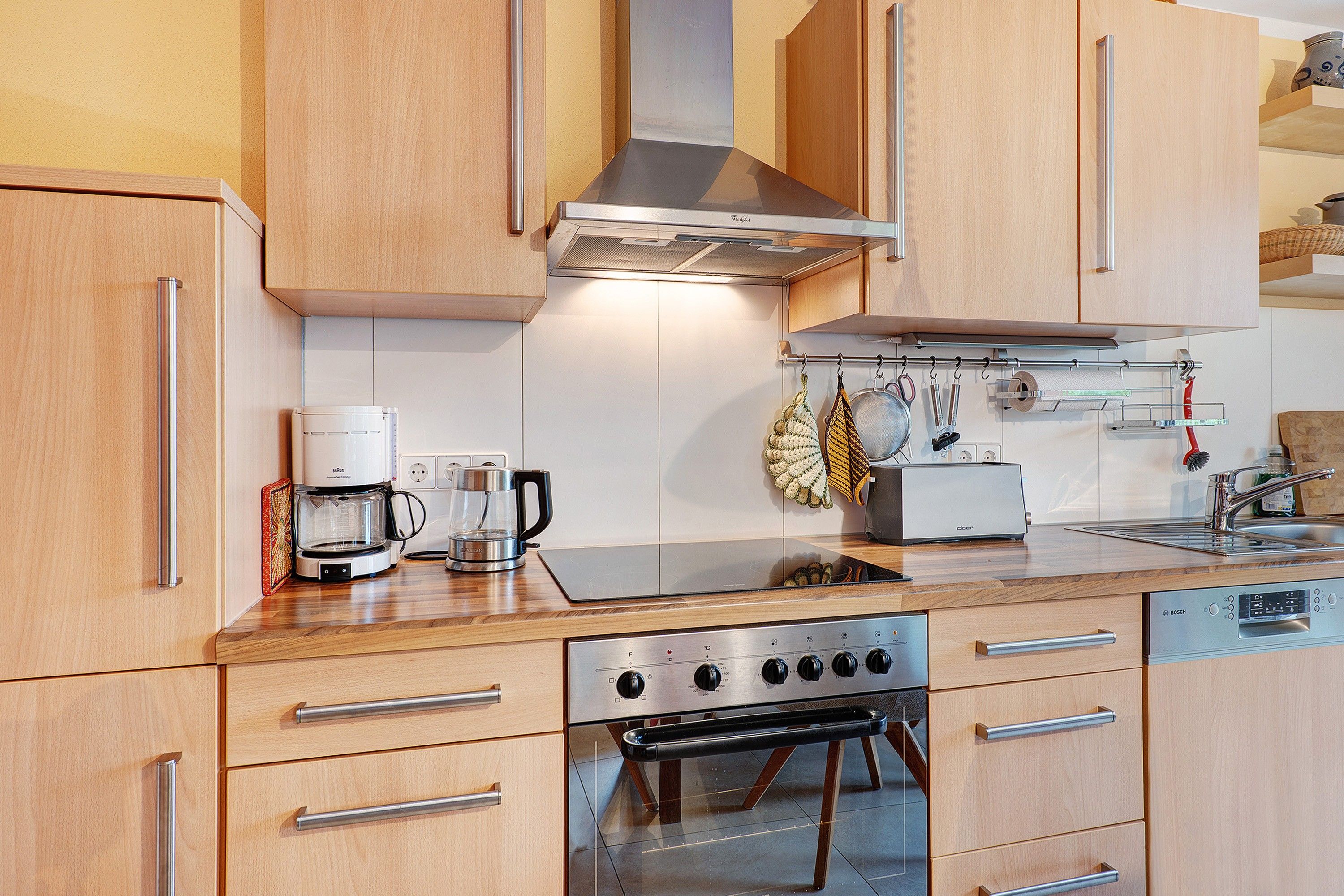 A modern kitchen with wooden cabinets, a stainless steel range hood, a coffee maker, a kettle, and a dishwasher.