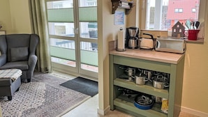 Una sala de estar acogedora con un sillón gris, un tapete estampado y una zona de cocina con cafetera, tostadora y utensilios de cocina en estantes.