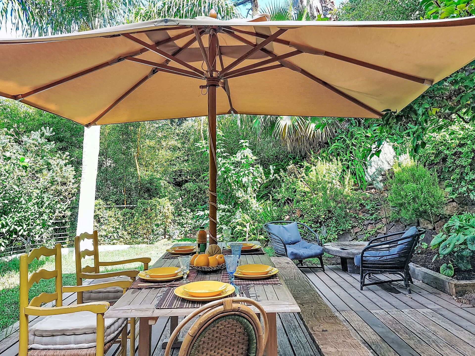 An outdoor patio with a wooden table, yellow chairs, a blue chair, and a large umbrella.