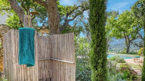 Une douche extérieure en bois, avec une serviette suspendue à une clôture en bois, entourée d’une végétation luxuriante et offrant une vue sur une piscine.