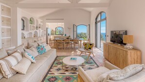 Un salon spacieux avec un grand canapé, une table basse et un coin repas avec des chaises. De grandes fenêtres offrent une vue sur l’océan.