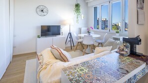 Un salon moderne avec un téléviseur, un coin repas avec des chaises, une table basse à motifs et une vue sur la ville à travers de grandes fenêtres.