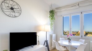 Un salon moderne doté d’un téléviseur à écran plat, de meubles blancs, d’une table à manger avec des chaises, d’un lampadaire et offrant une vue sur le paysage urbain grâce à de grandes fenêtres.